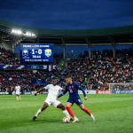 French player dribbling soccer ball past Ivorian defender in stadium with scoreboard showing France 1 Côte d'Ivoire 0